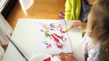 Genç bir sanatçı masadaki bir fırçayla oturmuş, parmaklarını ve tırnaklarını kullanarak eğlenceli ve renkli bir görsel sanat yaratıyor.