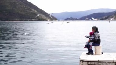 Anne kucağında küçük bir çocukla iskelede oturur ve denizin üzerinde uçan martılara bakar. Yüksek kalite 4k görüntü