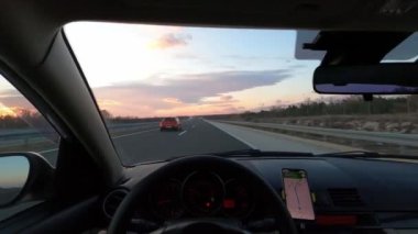 Bir motorlu araç gün batımında otoyolda ilerliyor, dikiz aynaları ve dikiz aynalarında renkli gökyüzünü ve bulutları yansıtıyor.
