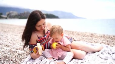 Küçük kız plajda annesinin yanında battaniyede otururken büyük bir elmayı kemiriyor. Yüksek kalite 4k görüntü