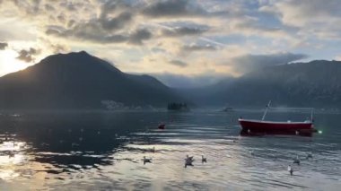 Kırmızı bir tekne huzurlu bir göl üzerinde süzülür. Görkemli dağlar, açık mavi bir gökyüzünün ve tüylü beyaz bulutların altında yükselir.