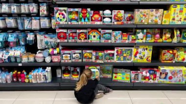 Bir perakende mağazasının zemininde oturan bir müşteri oyuncaklarla dolu bir rafta ürünleri inceliyor. Markette müşteriler için çeşitli ticaret yayınları sunulmaktadır.
