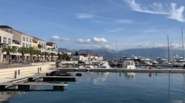 Huzurlu sulara kenetlenmiş teknelerle dolu, sakin bir liman. Arka planda, bulutlu, huzurlu bir gökyüzünün altında binalar ve evler çerçevelenmiş.