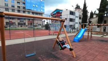 Bir çocuk, şehir çocuk parkında salıncakta sallanıyor, bina ve ağaçlardan oluşan bir arka planda. Elektrik mavisi gökyüzü sakin atmosferi güçlendirir