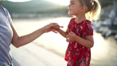 Bir kadın, parmakları birbirine dolanmış kırmızı elbiseli küçük bir kızın elini nazikçe tutar. Kızların elbisesinin kolsuz, tek kollu, yüksek bel ve su benzeri bir jesti var.