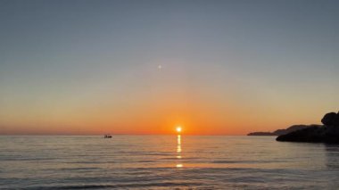 Güneş sıvı ufukta batarken, uzaklardan bir tekne görülebilir. Gökyüzü kırmızı ve turuncu renklerle boyanır, okyanus üzerinde güzel bir ışık saçar.