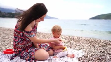 Anne ve küçük bir kız, sahildeki bir battaniyede oturmuş soyulmuş muz yiyorlar. Yüksek kalite 4k görüntü