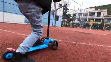 Kişi mavi bir scooter 'la pistte zumluyor, bacağı asfalttan itiyor. Tekerlekler zeminde pürüzsüzce dönüyor, güvenlik için kaskla korunuyor.