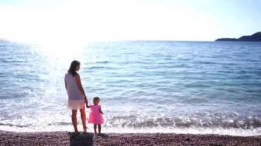 Annem ve küçük bir kız deniz kıyısında el ele tutuşuyorlar. Arkadan bak. Yüksek kalite 4k görüntü