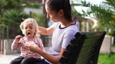 Küçük kız annesinin kollarında bankta otururken yemek kutusundan kaşıkla yulaf lapası yiyor. Yüksek kalite 4k görüntü