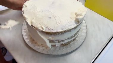 Bir insan masadaki spatulayı kullanarak pastaya krema sürüyor. Fırında pişirilen tatlı servis malzemesi ve bulaşık takımı ile hazırlanıyor.