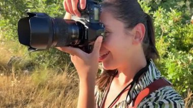 Bir kadın fotoğrafçı, güzel otlak bir alanda dijital bir kamerayla fotoğraf çekiyor. Doğayı keşfederken gülümsüyor ve fotoğraf sanatını icra ediyor.