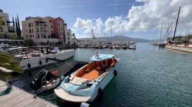 Bir su aracı, limanın yanında, bulutlu bir gökyüzünün altında demirlemiştir. Bina ve araçlar arka planda görülebiliyor ve seyahat ve deniz mimarisinin pitoresk bir sahnesini oluşturuyor.