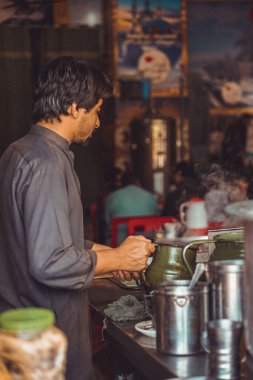 Pakistani man food vendor preparing smoking hot masala chai, the local tea, in a kettle in his roadside street food stall clipart