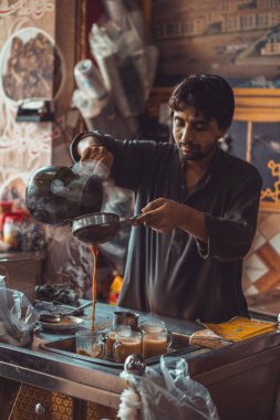 Pakistani man food vendor preparing and pouring smoking hot masala chai, the local tea, into cups from kettle in his roadside street food stall clipart