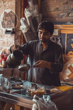 Pakistanlı erkek yiyecek satıcısı, yol kenarındaki yemek tezgahındaki çaydanlıktan gelen fincanlara sıcak masala çayı hazırlıyor ve döküyor.