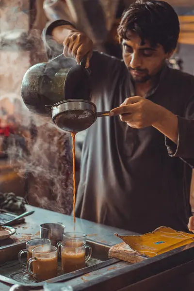 Pakistanlı erkek yiyecek satıcısı, yol kenarındaki yemek tezgahındaki çaydanlıktan gelen fincanlara sıcak masala çayı hazırlıyor ve döküyor.