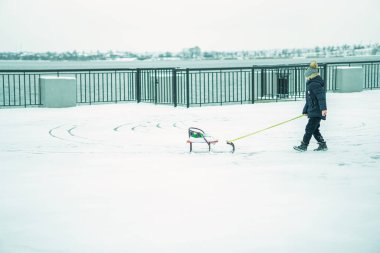 Bir çocuk kızağıyla karda oynuyor. Nehir kenarında karla kaplı bir sokak..