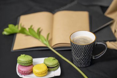 eskiz defteri, defterler, bir fincan kahve, beyaz tabakta renkli badem kurabiyeleri, siyah arka planda yeşil bir dal..