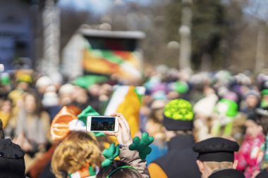 Kalabalıktan oluşan bulanık bir arka plan. Cep telefonunda yeniden çekilme fotoğrafı. Aziz Patrick Günü karnavalından elinde akıllı telefon videosu olan bir kızın elleri