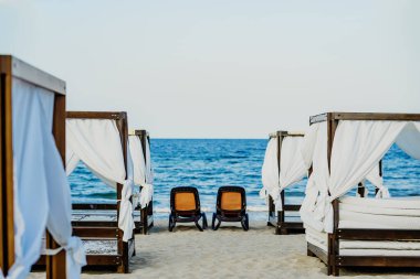 Bir dizi beyaz kabin kumlu bir plajda durup güneşten uzak durmayı teklif ediyor. Gök mavisi sular arka planda uzanıyor, rahatlama ve sükuneti davet ediyor.