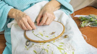 İşlemeli bir işte çalışan bir kadının elleri kumaşa çiçekli bir desen dikiyor. Karmaşık iğneleme sanatını vurguluyor.