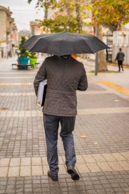 Siyah şemsiyeli bir adamın ıslak bir şehir sokağında yürüdüğünü kolunun altında belgeleri tuttuğunu hayal edin.