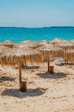 Arka planda turkuaz deniz bulunan kumlu bir sahilde saman şemsiyeler. Egzotik tatil beldesi atmosferli tropikal yaz inzivası. Saman şemsiyeli sakin bir sahil, turkuaz deniz kenarında güneşli bir gün.