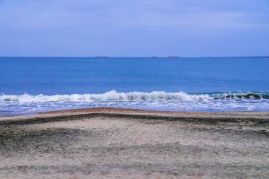 Dalgalar deniz kabuğu kaplı kumsala çarpar. Mavi gökyüzünün altında doğal siyah kum ve ufukta kargo gemileri vardır. Karadeniz kıyısında dalgalar, Burgaz Körfezi, Bulgaristan. Seyahat kavramı