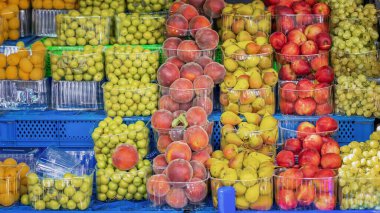 Yerel markette renkli taze meyveler, şeftaliler, erikler, elmalar ve üzümler açık kaplarda. Plastik tepsilerdeki meyveler yerel üretim çeşitliliğini vurguluyor