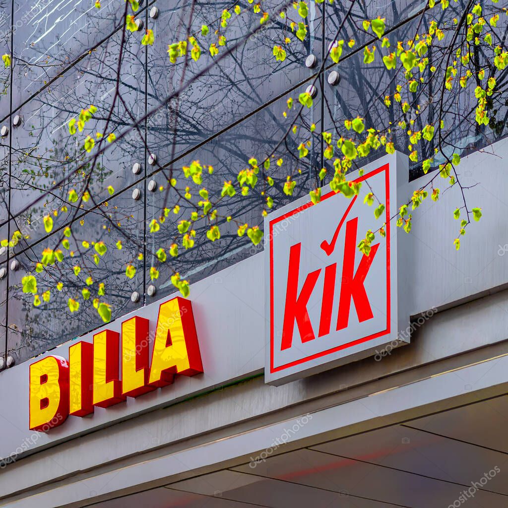 Storefront signs of Billa and KiK captured on rainy day with spring leaves in foreground. Store signs highlight retail branding, urban presence and, commercial identity