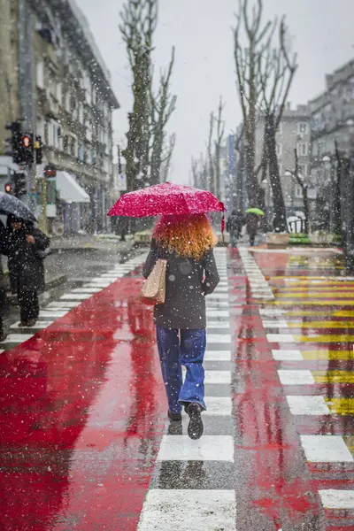 Kıvırcık kızıl saçlı kadın ıslak şehir yaya geçidinde kar altında parlak kırmızı şemsiye altında yürüyor. Mevsimler kavramı, yağmurlu günler, şehir yaşam tarzı, kış havası, şehir yalnızlığı