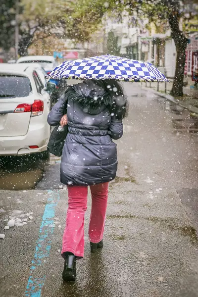 Kar yağarken şemsiyeyle koşuşturan ve ıslak şehir caddesinde yağmur yağan bir kadın. Kentsel kış havası, samimi sokak fotoğrafı. Mevsimler, hava durumu