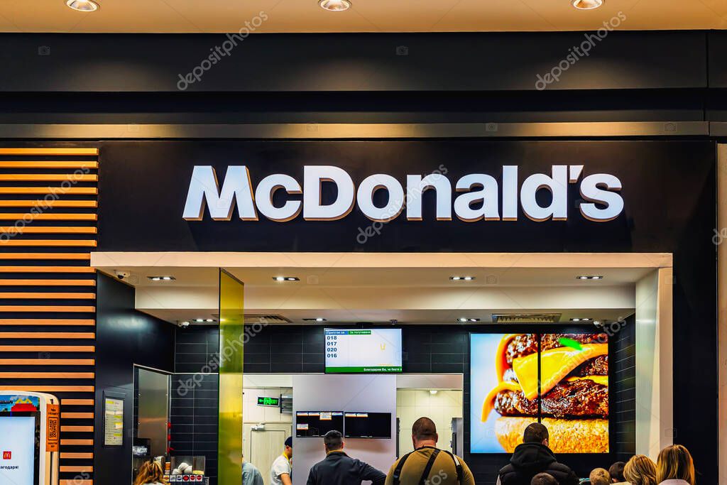 McDonalds restaurant interior with illuminated logo, digital menus, and people queuing for food in food court. Concept of McDonalds branding, fast food culture, global identity