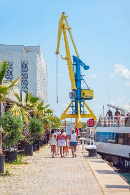 Güneşli liman, Burgaz limanı, Bulgaristan. Turistler rıhtım, rıhtım, parlak vinç ve rıhtıma yanaşmış tekneler boyunca yürüyorlar. Sahil turizmi, yaz seyahati, şehir yaşam tarzı kavramı