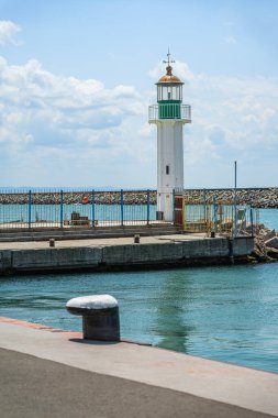 Yeşil aksanlı eski beyaz deniz feneri rıhtıma yakın, parlak gün ışığında limanda çitlerle çevrili. Deniz feneri kavramı, sahil güvenliği, navigasyon yapısı