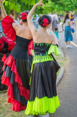 Kırmızı çiçekli canlı elbiseler içinde flamenko dansçıları parktaki açık hava festivalinde geleneksel hareketlerle şehir parkında yürüyorlar. Flamenko konsepti, kültürel performans, İspanyol Kültür Festivali