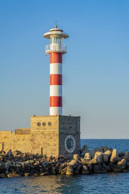 Kırmızı ve beyaz deniz feneri, sakin deniz kıyısında, akşam güneşinin altında kayalık dalgalı sularda dimdik ayakta duruyor. Deniz feneri kavramı, denizcilik sembolü, denizcilik navigasyonu, kıyı manzarası ve güvenli deniz yolculuğu.