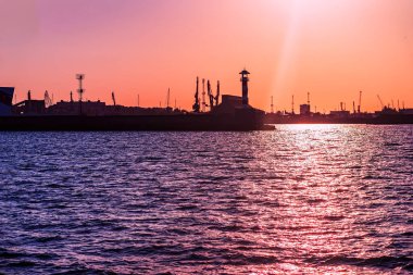 Canlı turuncu günbatımı, denize yansıyan vinçler ve çizgili deniz fenerleriyle iskele ufuk çizgisini aydınlatıyor. Deniz feneri kavramı, sanayi kıyıları, renkli deniz endüstrisi geçmişi