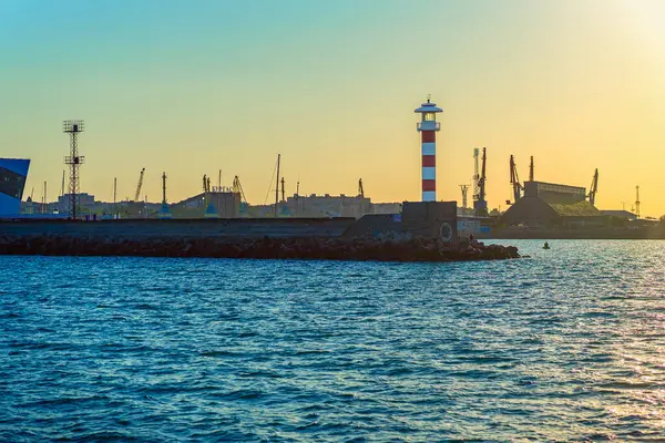 Gün batımında deniz feneri olan sanayi limanı. Deniz feneri endüstriyel limanın kenarında duruyor, vinçlerle, altın günbatımı gökyüzü. Deniz feneri kavramı, şehir limanı, doğa ve altyapı arasındaki zıtlık