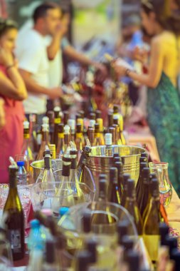 Şarap festivali, şişeler ve insanlar, bulanık arka plan. Bir sürü şarap şişesi, buz kovası, tatma masası, sosyalleşen insanlar, arka planda örnekler. Şarap tatma, kutlama, sosyal etkinlik kavramı.