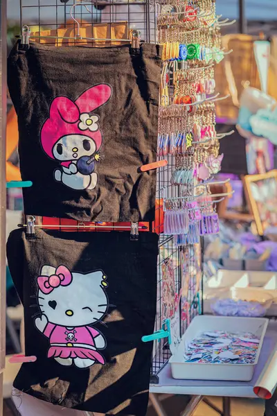 Merhaba Kitty Poşetleri ve Anahtarlıkları. İçinde Hello Kitty ve Melody tasarımlarım olan sevimli siyah çantalar renkli anahtarlıkların yanında sergileniyor. Kawaii modası, şirin aksesuarlar, pop ikonu ürünleri.