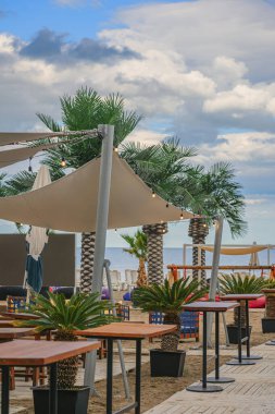 Palm Trees 'li açık hava plaj kafesi, ahşap masalar ve kumlu sahilde bulutlu gökyüzü olan dekoratif ışıklar. Plaj kafe kavramı, tropikal deniz kenarı rahatlığı, yaz tatili.
