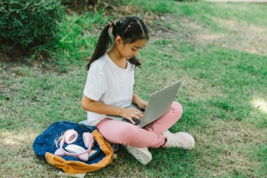 Parktaki çimlerde oturan ve dizüstü bilgisayar kullanan tatlı küçük kız.