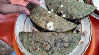 Bir el, peynir ve sebzeyle doldurulmuş mavi mısır tortillasıyla yapılan geleneksel bir Meksika yemeğinin olduğu tabağa uzanır. Tabağın canlı renkleri mutfağın kültürel zenginliğini vurguluyor..