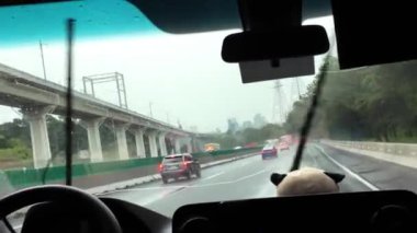 Düşük görüş mesafesi ve nemli koşullarda yağmurlu bir yolda bir araç yol alır. Her iki taraftan da arabalar geçerken arka planda uzun binalar görülebilir..