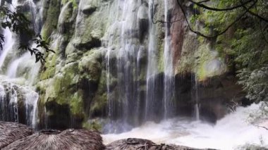 Güzel bir şelale, tropik bir çevrede yemyeşil yemyeşil kayaların üzerine yuvarlanır. Geleneksel sazdan kulübeler yakınlardayken, akan suyun sesi doğanın sükunetiyle harmanlanır..