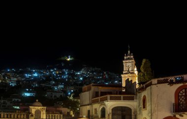 Taxco de Alarcon showcases its rich heritage at night, with the hills adorned by twinkling lights from the homes. The skyline features historical buildings, including a church with a striking clock tower. clipart