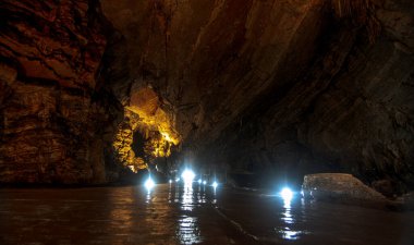 Visitors explore the stunning Grutas de Cacahuamilpa National Park, surrounded by impressive rock formations and shimmering waters. The atmosphere is illuminated by strategically placed lights that highlight the beauty of the cave walls. clipart