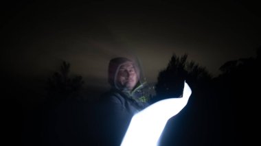 Lone figure exploring a dark forest at night with bright flashlight clipart